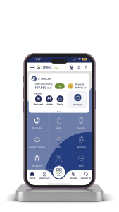 A 3D mockup of a dark purple smartphone standing on a brushed metal base. The screen displays the "ONEIC Pay" mobile application, a digital wallet and utility payment app. The interface features a "Total Outstanding" balance of 407.580 OMR, quick action buttons for "Merchant," "Mobile," and "TopUp," and a grid of service icons including Electricity, Water, Telecom, and Insurance.